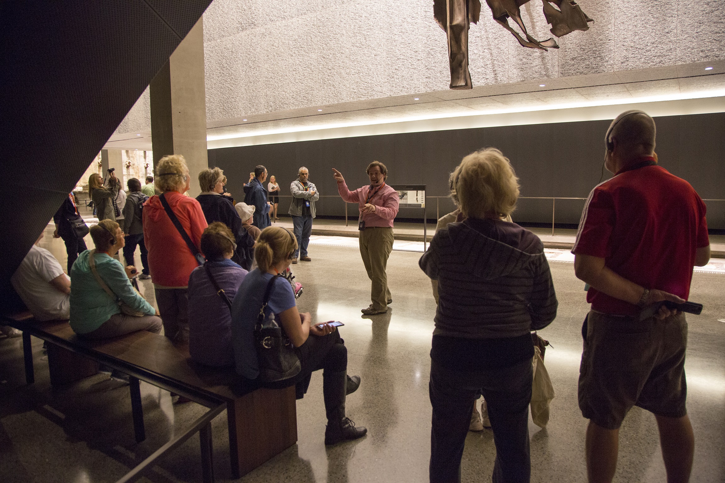 A guide leads a tour inside the 9/11 Memorial & Museum. About a dozen people watch on as he talks and gestures at an object unseen.