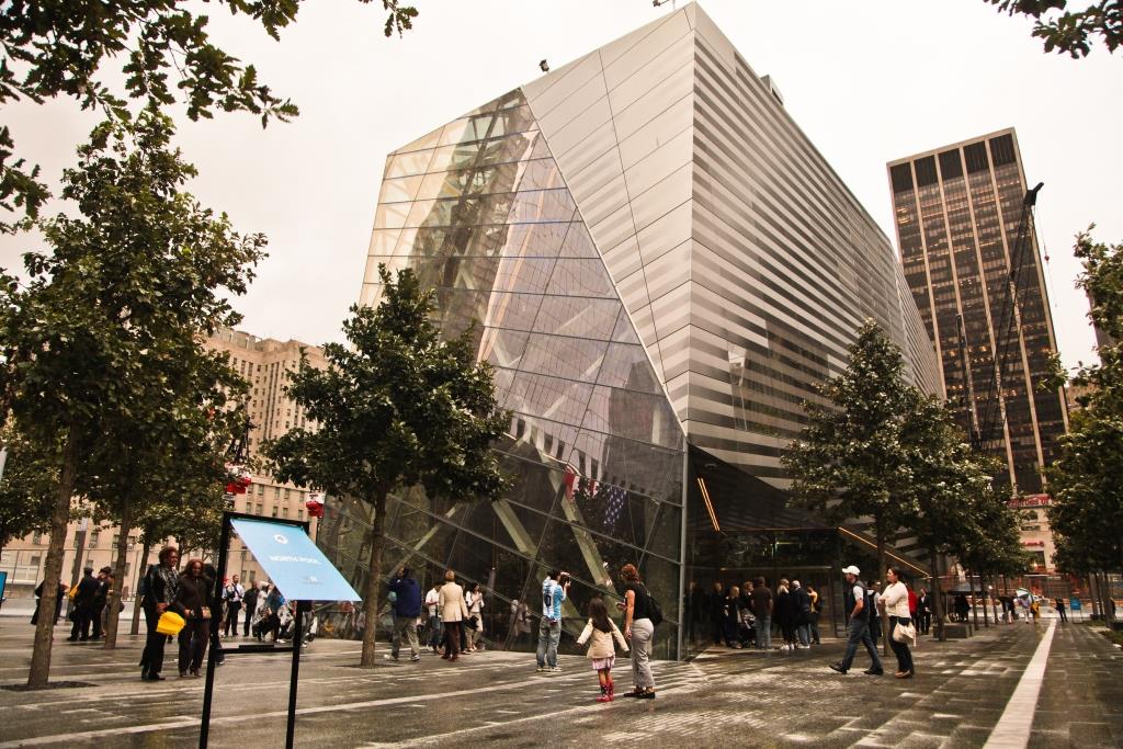 Dozens of visitors walk around the glass and steel facade of the Museum pavilion on Memorial plaza. 