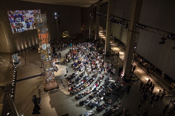 Dozens of seated people watch several people speaking onstage beside the Last Column at Foundation Hall. A video is projected on a wall and the lighting in the hall has been dimmed for the event.