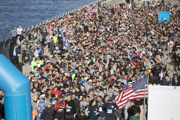 Hundreds of participants gather for the 9/11 Memorial & Museum 5K Run/Walk and Community Day. A large American flag is in the foreground. 