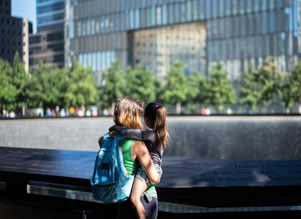 A woman with a backpack holds a young girl with a ponytail as they both look at one of the Memorial’s reflecting pools on a sunny day. Green oak trees and buildings are seen farther afield.