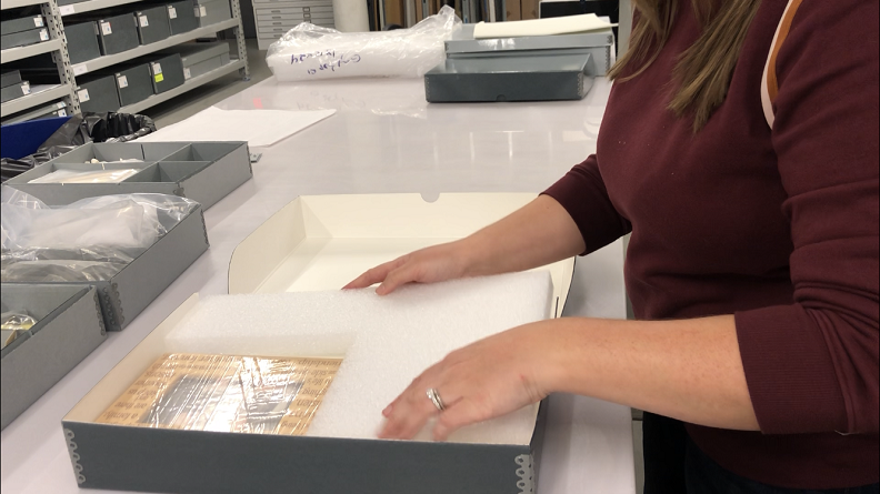 A woman in a burgundy sweater packages an artifact in Styrofoam in a gray box.