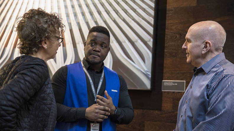 A volunteer wearing a distinctive blue vest listens to two visitors, a man and a woman.