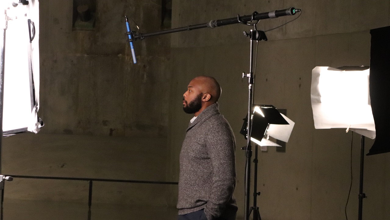 In this photograph, a man stands in profile before a boom mic and set lights during the Anniversary in the Schools webinar filming.