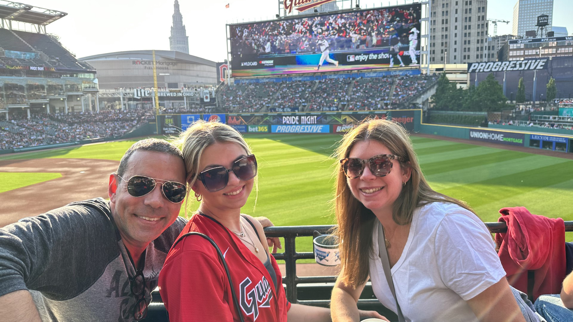 Three smiling people sitting in the stands of a baseball game.