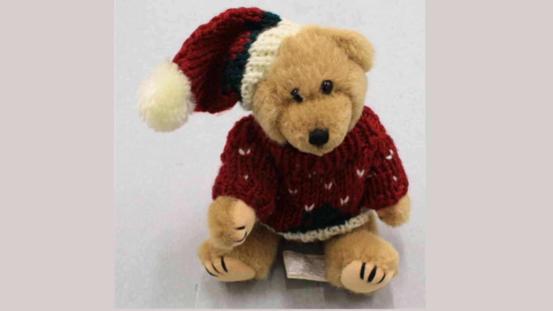 A light brown teddy bear wearing a maroon holiday sweater and matching Santa hat sits on a gray background