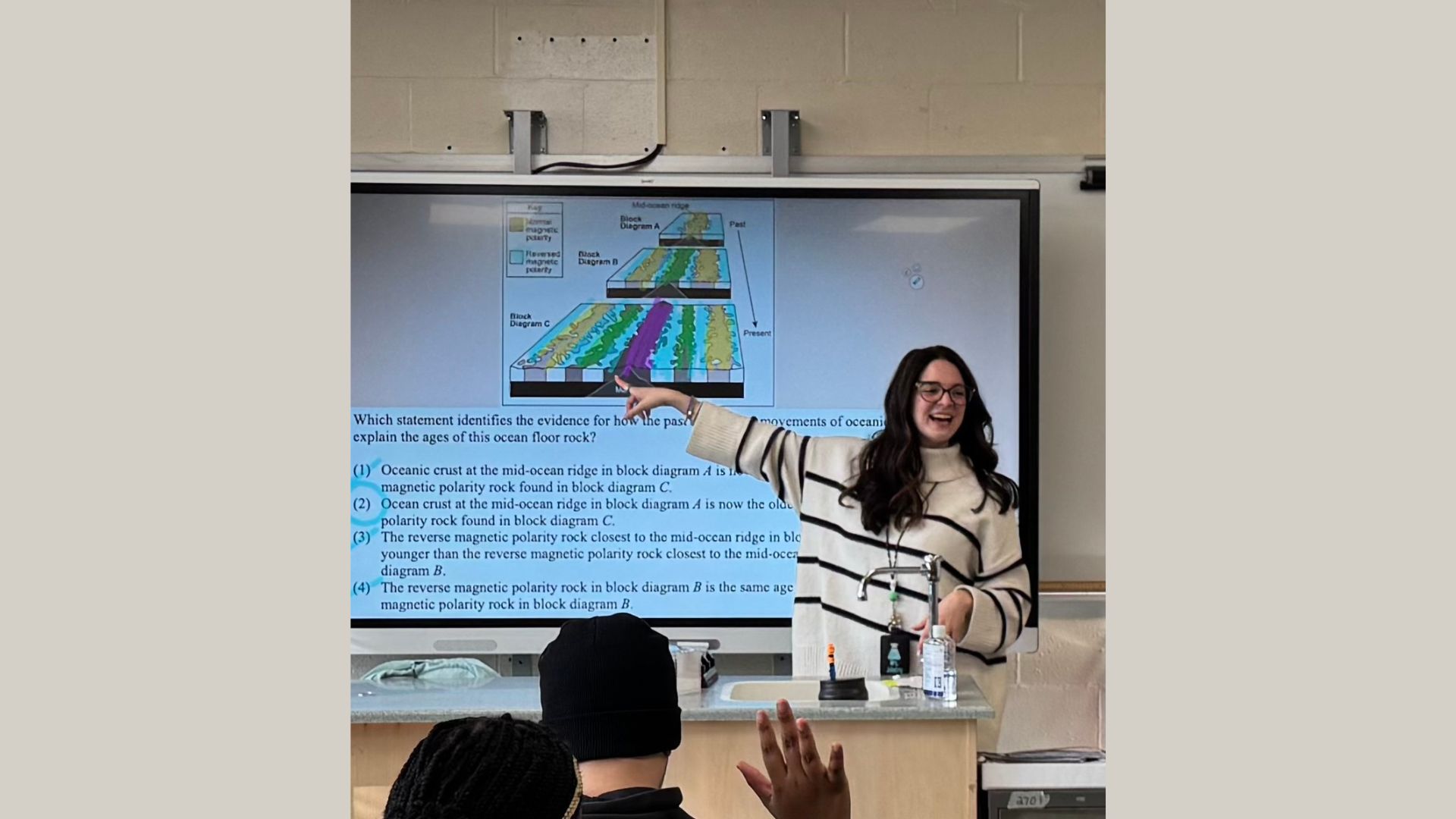 A teacher stands in front of her students, pointing to a presentation on screen