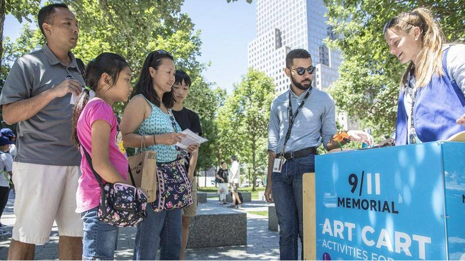 A small group of adults and children stand in front of the Art Cart, engaging with two Museum staff members 