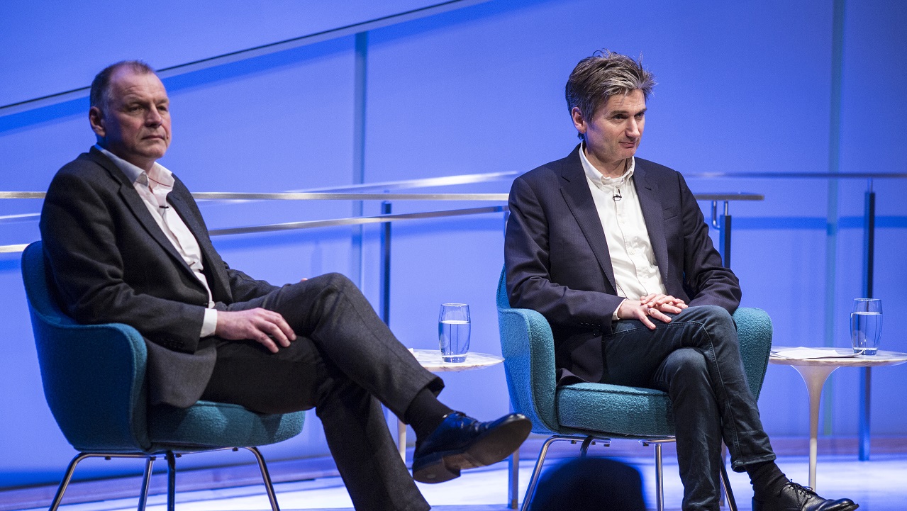 Two men sit with crossed legs on a blue-lit auditorium stage during a 9/11 Memorial Museum public program.