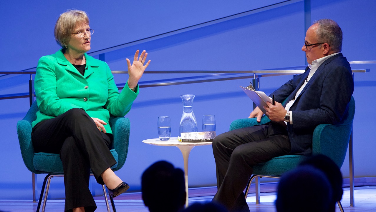 A woman in a green suit jacket speaks with her hands directed at a male moderator, who listens thoughtfully. The heads of audience members appear in silhouette in the foreground.