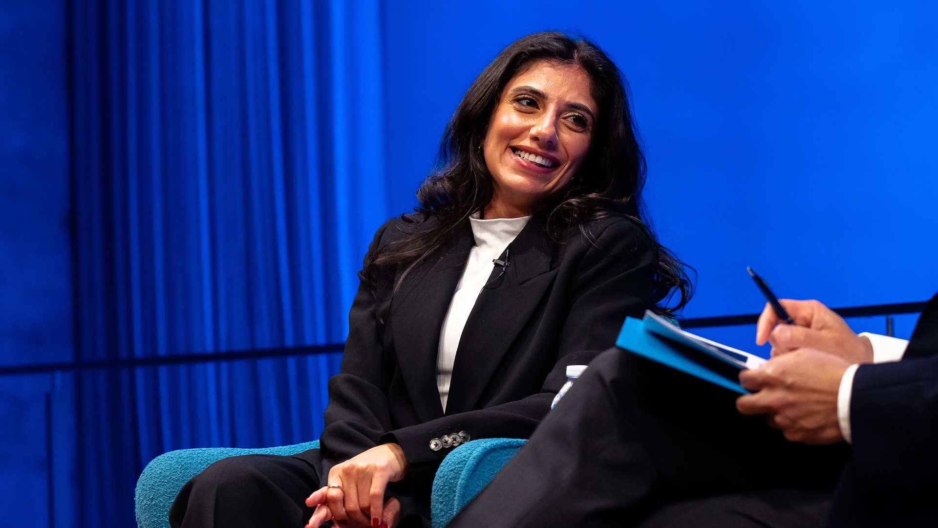 A woman with long, dark hair sits on stage, smiling, with a blue curtain in the background.