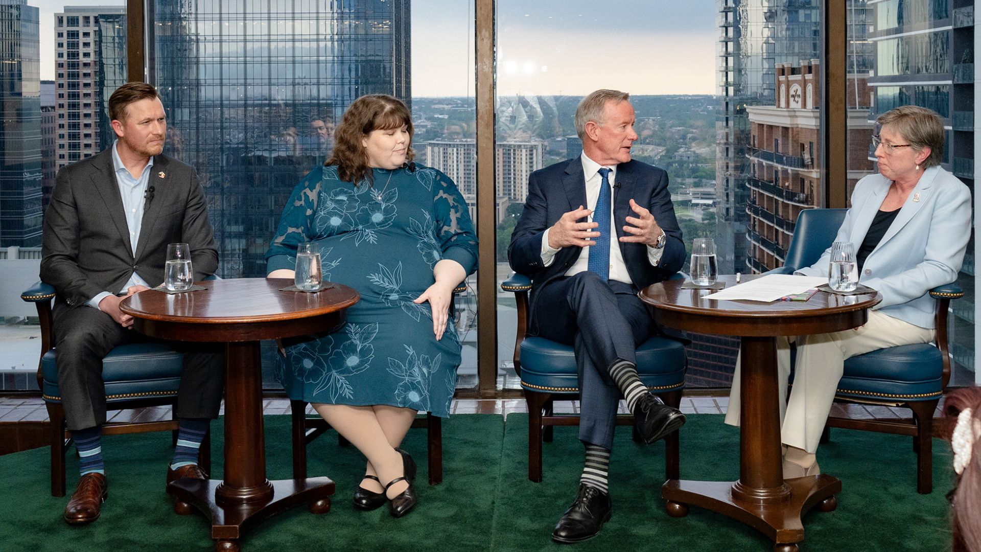 Four speakers, two men and two women, seated in front of a view of the city out of the window behind them