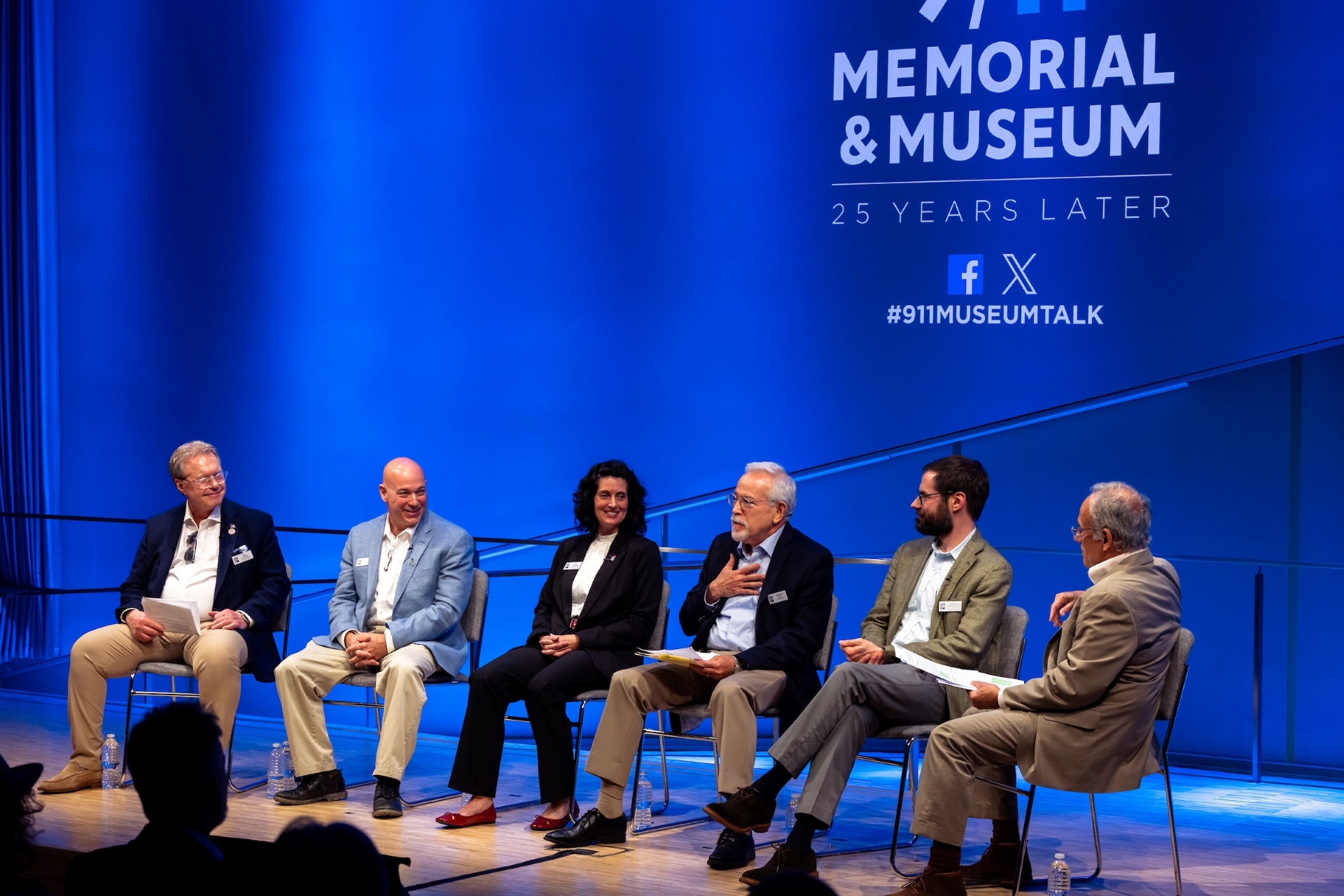 the group of six people sit on stage in front of a blue 9/11 memorial and museum background