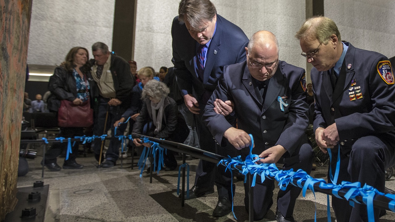 Several members of the FDNY and the family members place blue ribbons on a black railing at the foot of the Last Column.