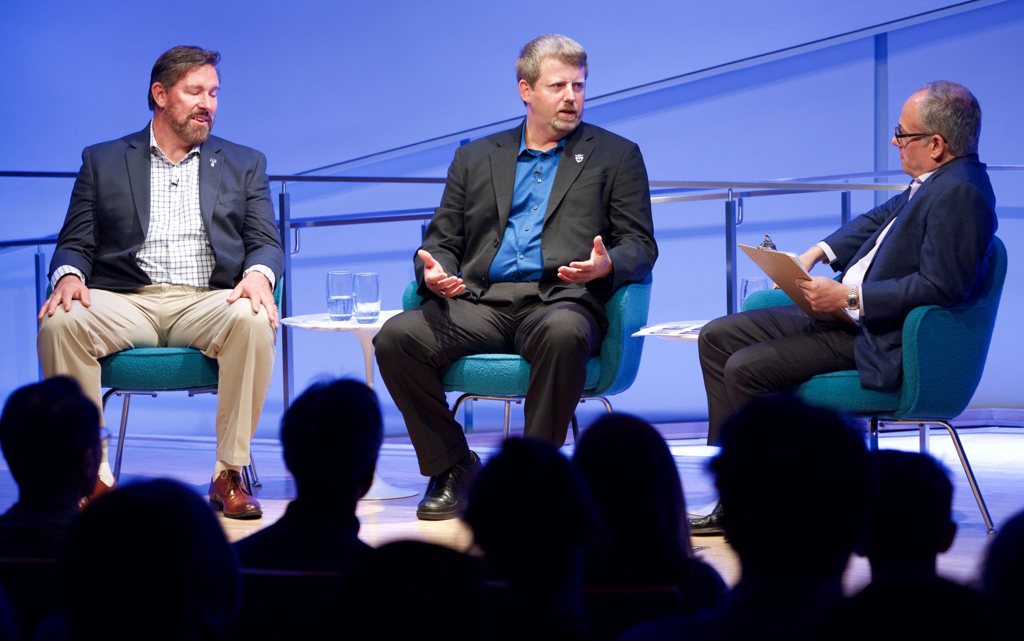 Former special forces commanders Maj. Mark Nutsch and retired Master Sgt. Scott Neil speak onstage as they take part in the public program, The Horse Soldiers. Clifford Chanin, the executive vice president and deputy director for museum programs, is seated to their left holding a clipboard. Audience members in the foreground are silhouetted by the stage lights. 