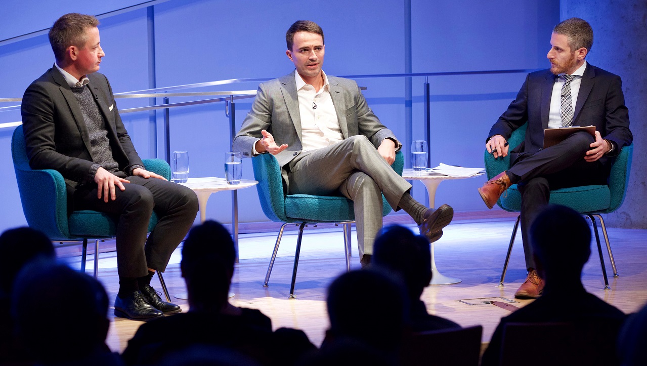 Three men in suits sit on a blue-lit auditorium stage. The man in the center gestures with his hands as the two men on either side of him look on and listen. The heads of the audience members appear in the foreground in silhouette.