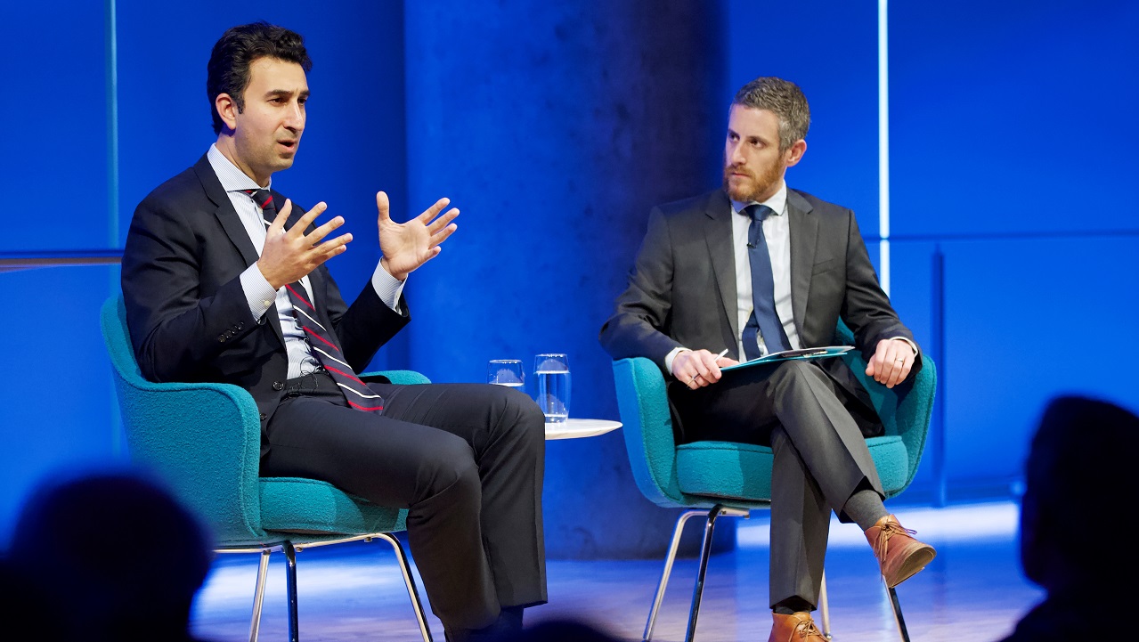 Two men in suits, a public program participant and a moderator, sit on a blue-lit auditorium stage. The program participant gestures with his hands while speaking out to the audience, whose heads appear in silhouette. The moderator looks on.