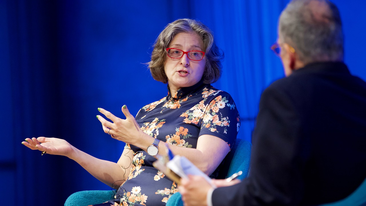 A public program participant sits on a blue-lit auditorium stage and gestures with her hands. The program moderator sits and listens with his back to the camera.