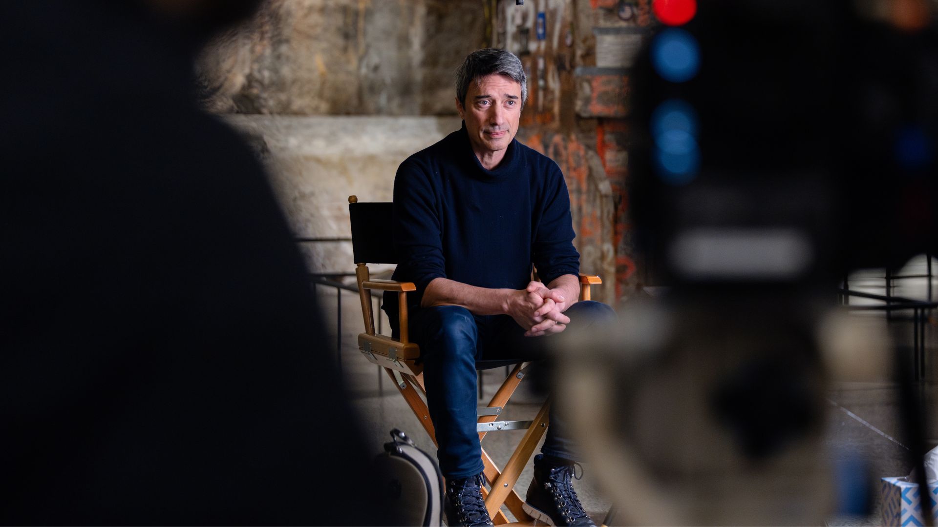 A man with graying hair sits in a director's chair, hands crossed, with the Last Column in the background. he is wearing jeans and a dark blue sweater.