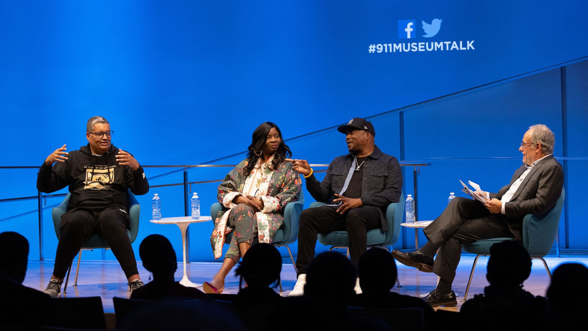 Panelists on stage at the 9/11 Memorial Museum. A bright blue screen sits behind them, with the social media icons for Twitter and Facebook and the text hashtag 911 Ed Talk superimposed on it.   