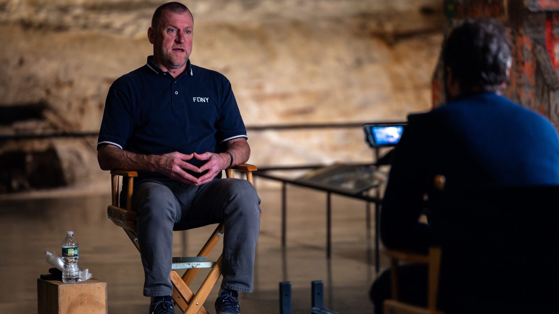 A person in a navy blue polo shirt - monogrammed with "FDNY" - sits in a director's chair facing an interviewer, with the Slurry Wall visible in the background.