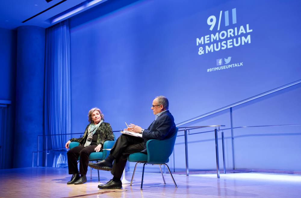 Dr. Sheila Carapico, professor of political science and international studies at the University of Richmond, sits onstage in this view taken from the stage. She is seated next to Clifford Chanin, the executive vice president and deputy director for museum programs, who is speaking as he holds a clipboard. Blue and white lights shine on the stage and on the walls behind it.