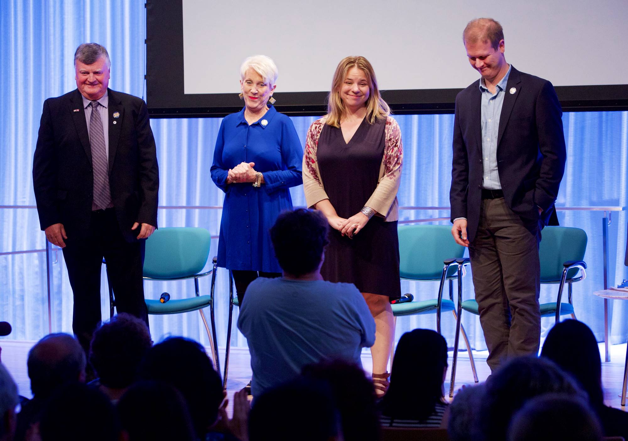 Four behind the award-winning new musical “Come From Away” stand onstage and look towards members of the audience as they smile. A member of the audience is standing and addressing them with a microphone in the foreground. Other audience members beside her are silhouetted by the lights of the stage.