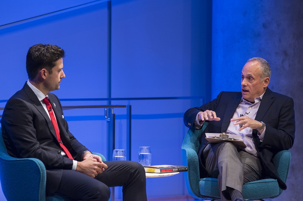 Two men sit on a blue-lit auditorium stage. One man in a suit and red tie sits and listens to a man with crossed legs and a clipboard in his lap while he speaks.