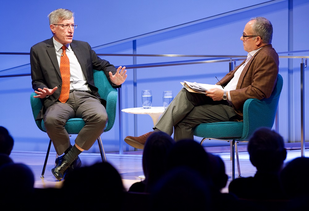 Two men sit on a blue auditorium stage. One man in a suit and orange tie gestures with his hands as he speaks. The other man sits with a clipboard in his lap and listens. The heads of the audience members appear in silhouette in the foreground of the photograph.