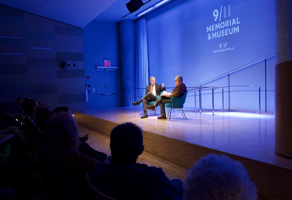 In this wide-angle photograph, two men sit on a blue auditorium stage while several rows of audience members look on.