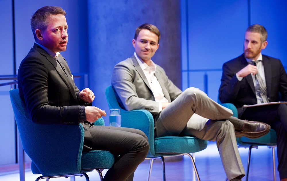 Three men in suits sit on a blue-lit auditorium stage. A man in a charcoal gray suit addresses an unseen audience while the other two men listen.