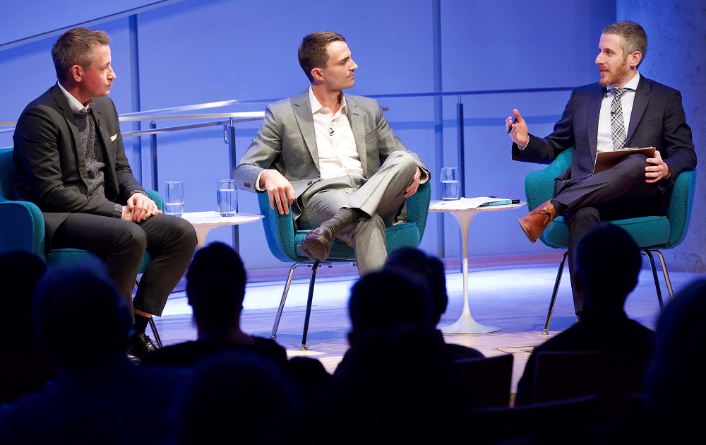 Three men in suits sit on a blue-lit auditorium stage. A man with a clipboard in his lap gestures with his hands toward the other two men, who sit with expressions of patient listening. The heads of the audience members appear in the foreground in silhouette.