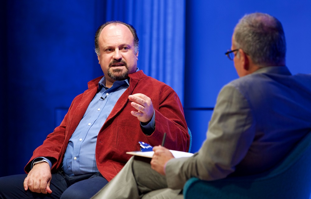 Two men sit on a blue-lit auditorium stage. One faces the camera and gestures with his hands.