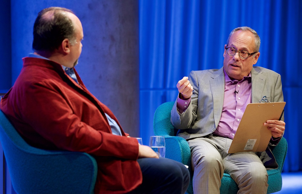 Two men sit on a blue-lit auditorium stage. One faces the camera and speaks while holding a clipboard.