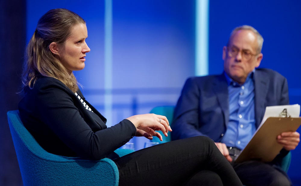 A female speaker appears in profile and gestures with her hands on a blue-lit auditorium stage while the male moderator looks on.