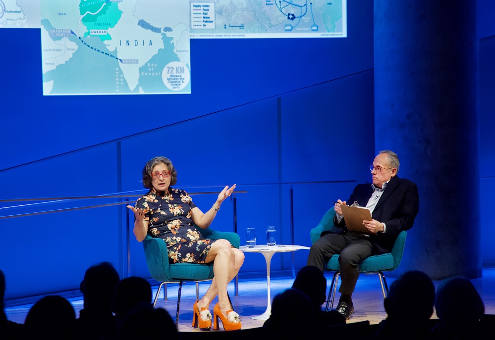 A public program participant sits on a blue-lit auditorium stage and gestures with her hands while looking out into the audience, whose heads appear in the foreground in silhouette. The program moderator sits to her left and looks on. Maps of India and Pakistan are projected onto the wall behind them.