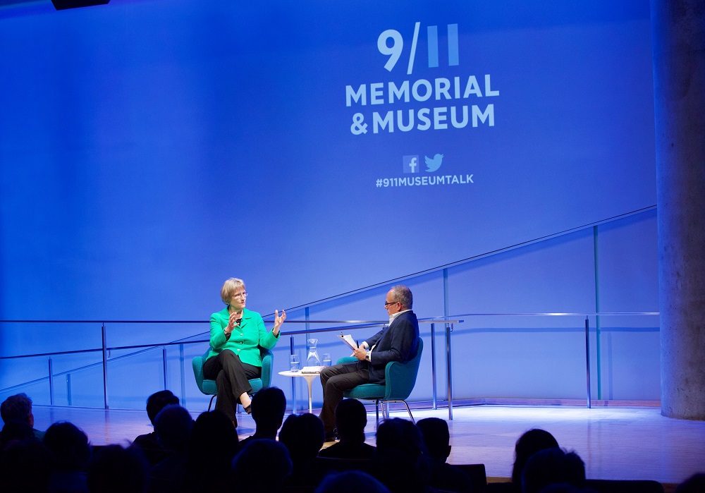 In this wide-angled photograph, A woman in a green suit jacket speaks on a blue-lit auditorium stage to a moderator. The heads of audience members appear in silhouette in the foreground.