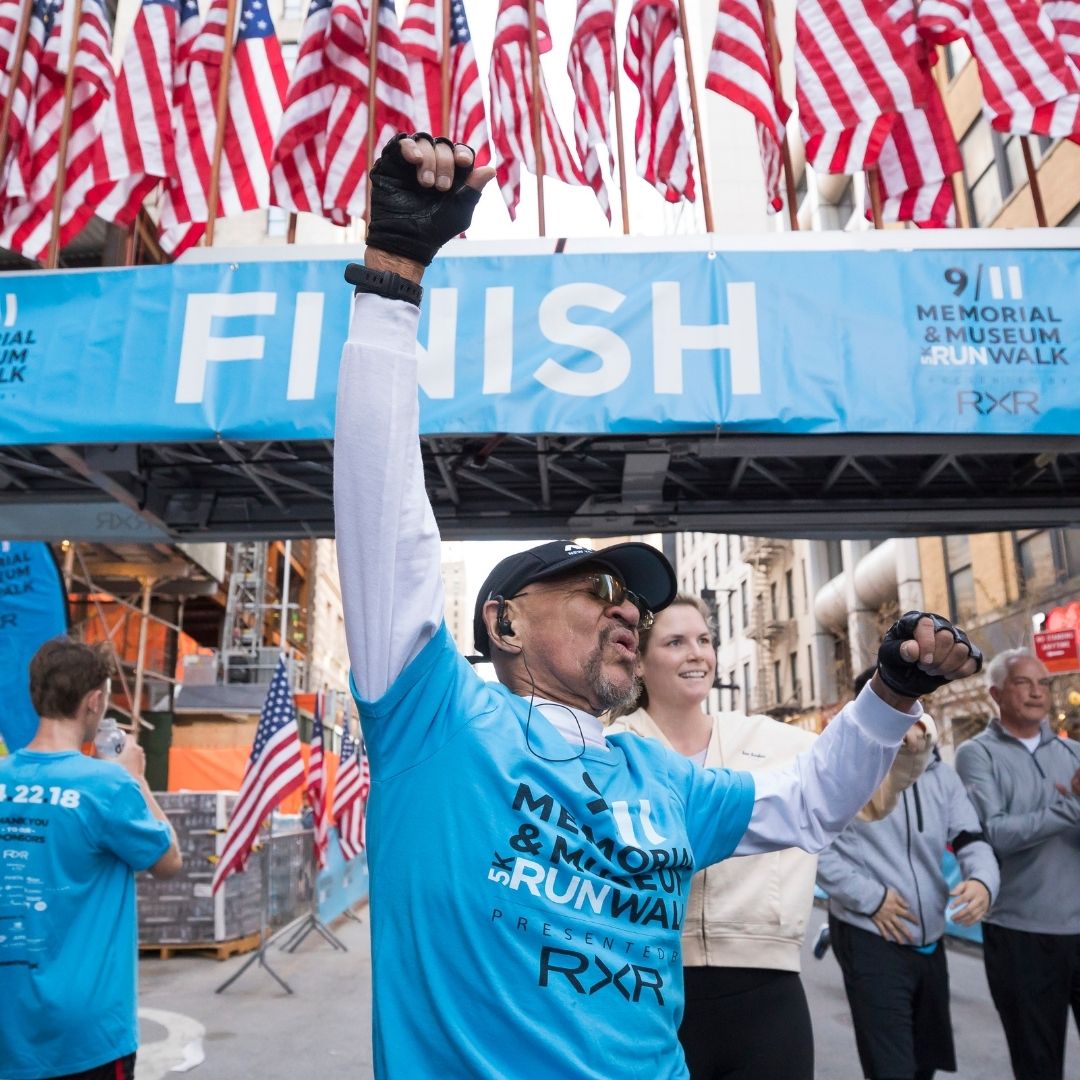 A man in a blue short-sleeve t-shirt over a white long-sleeve t-shirt crosses the finish line with one arm raised in victory