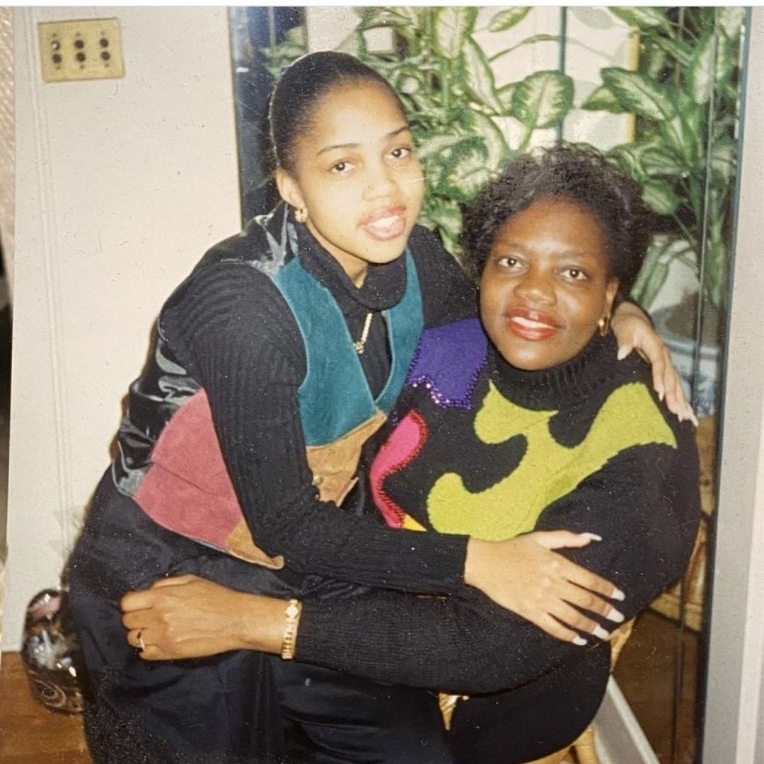 A younger dark-skinned woman sits on an older woman's lap. On the left, the younger woman wears a black, teal, mustard, and pink top. The woman on the right wears yellow and black.