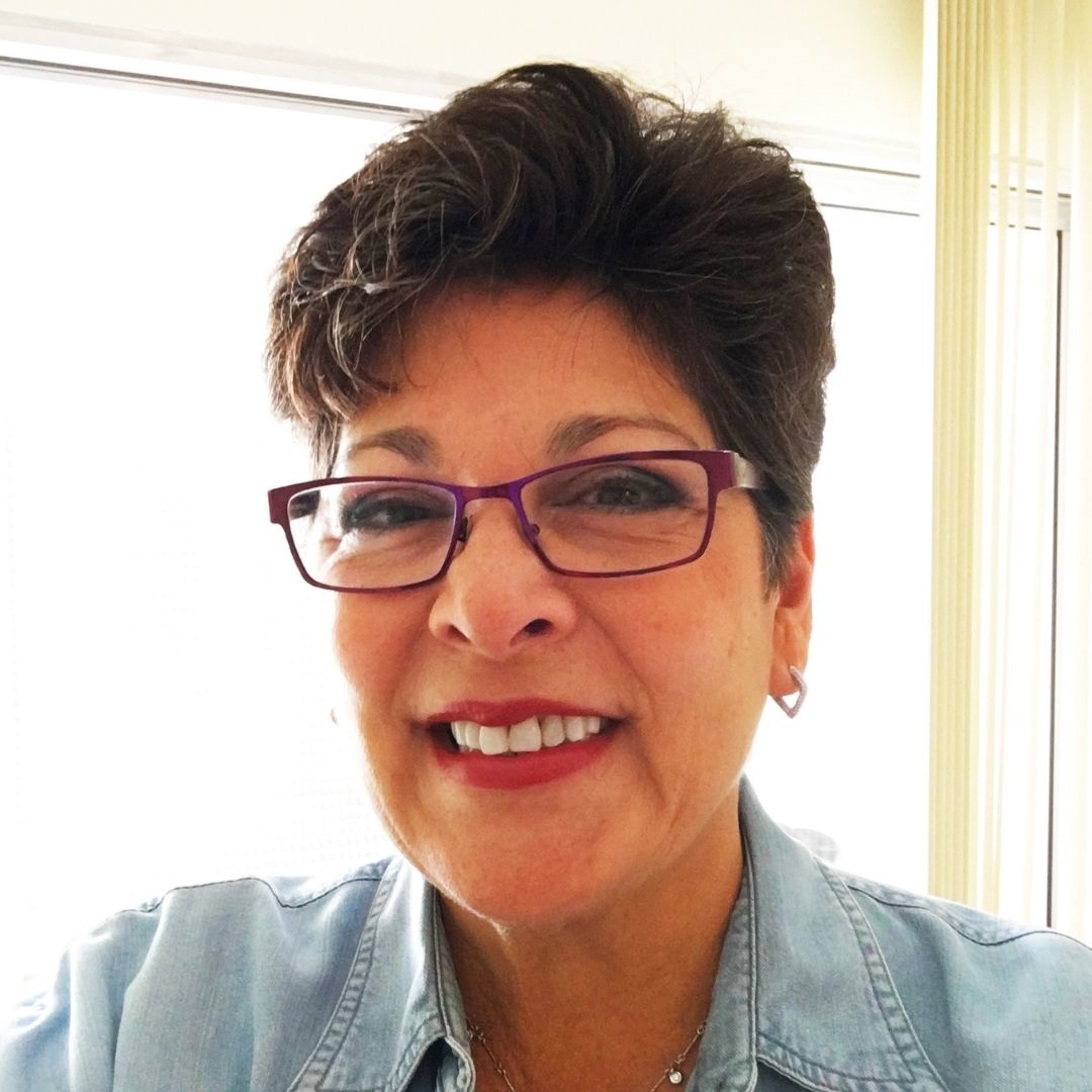 A woman with short, dark hair and red glasses, wearing a light blue shirt, smiles.