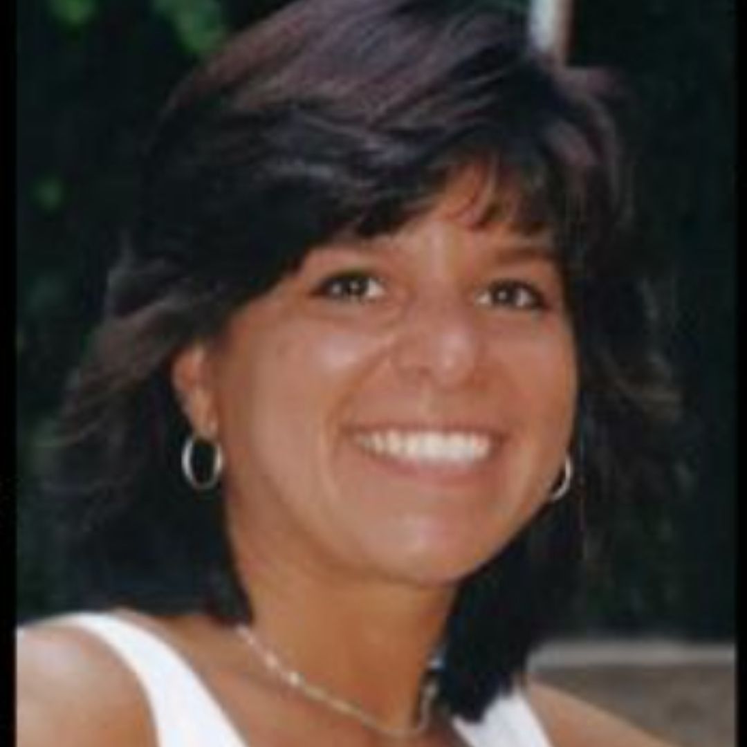 A woman with mid-length dark hair, wearing a white tank top and small hoop earrings, smiles. 