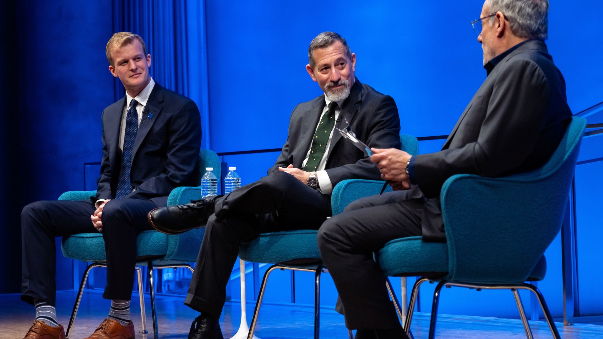 Three men on stage, all wearing dark suits, against a blue background displaying the 9/11 Memorial and Museum logo.