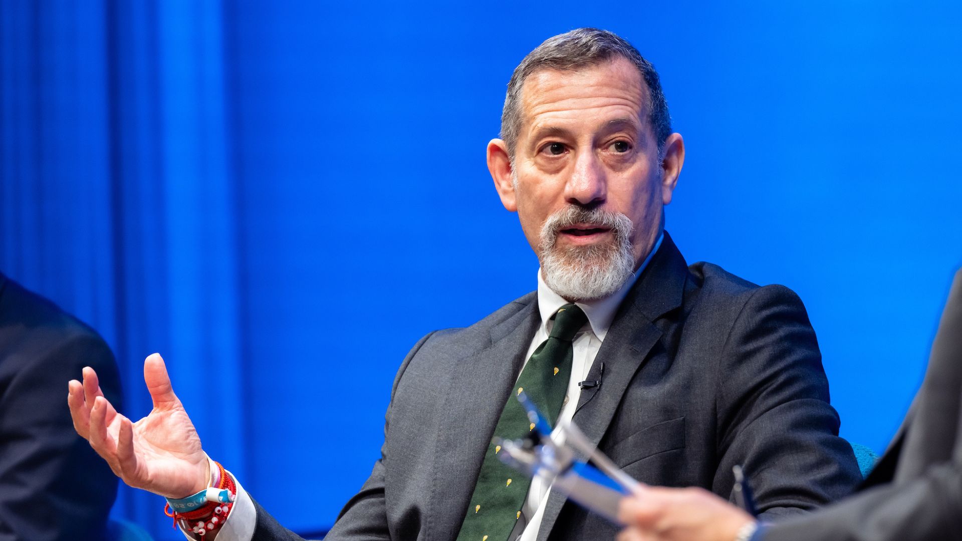 A man in a dark suit with a graying beard speaks on stage in front of a bright blue curtain.