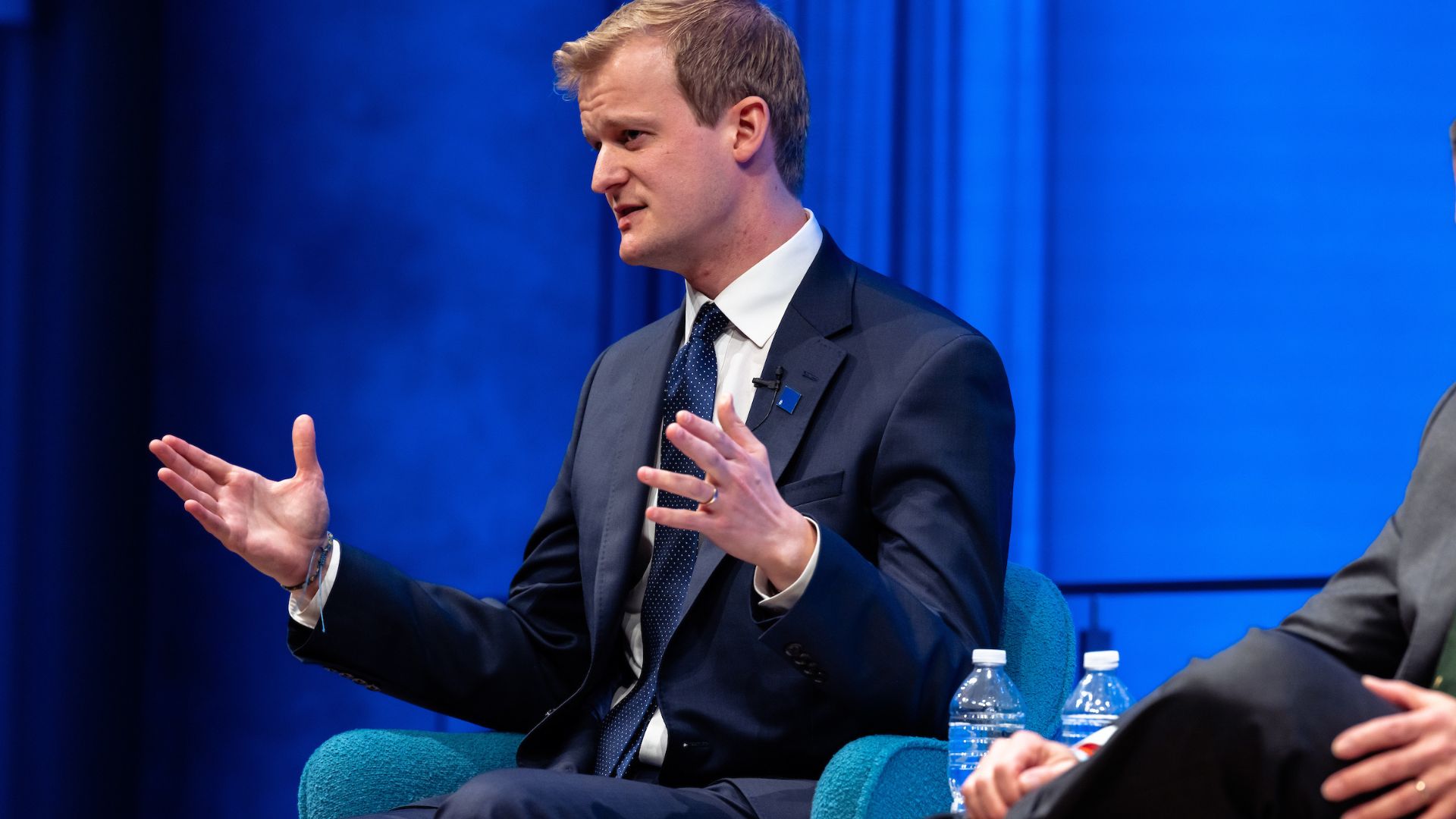 A blonde man in a dark suit speaks on stage in front of a bright blue curtain.