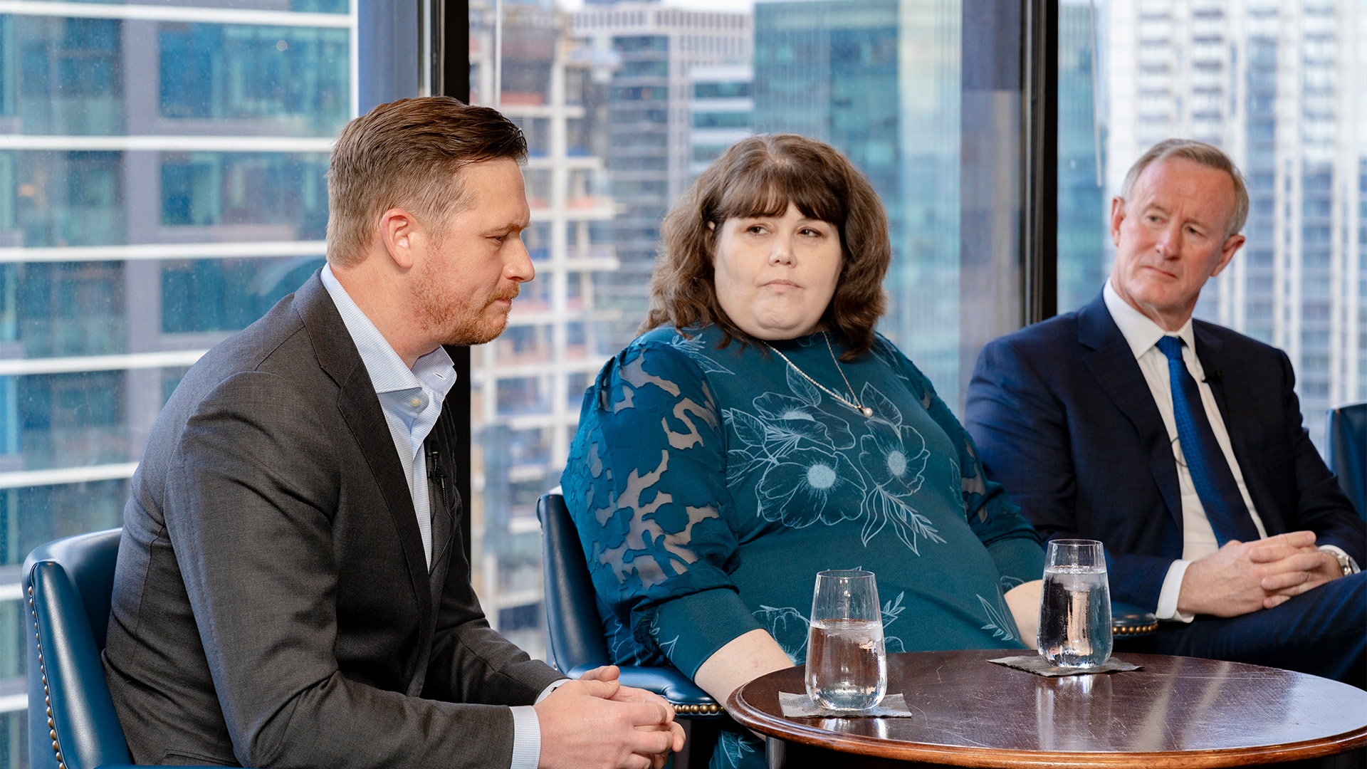 Three speakers, one woman and two men, seated in front of a view of the city out of the window behind them