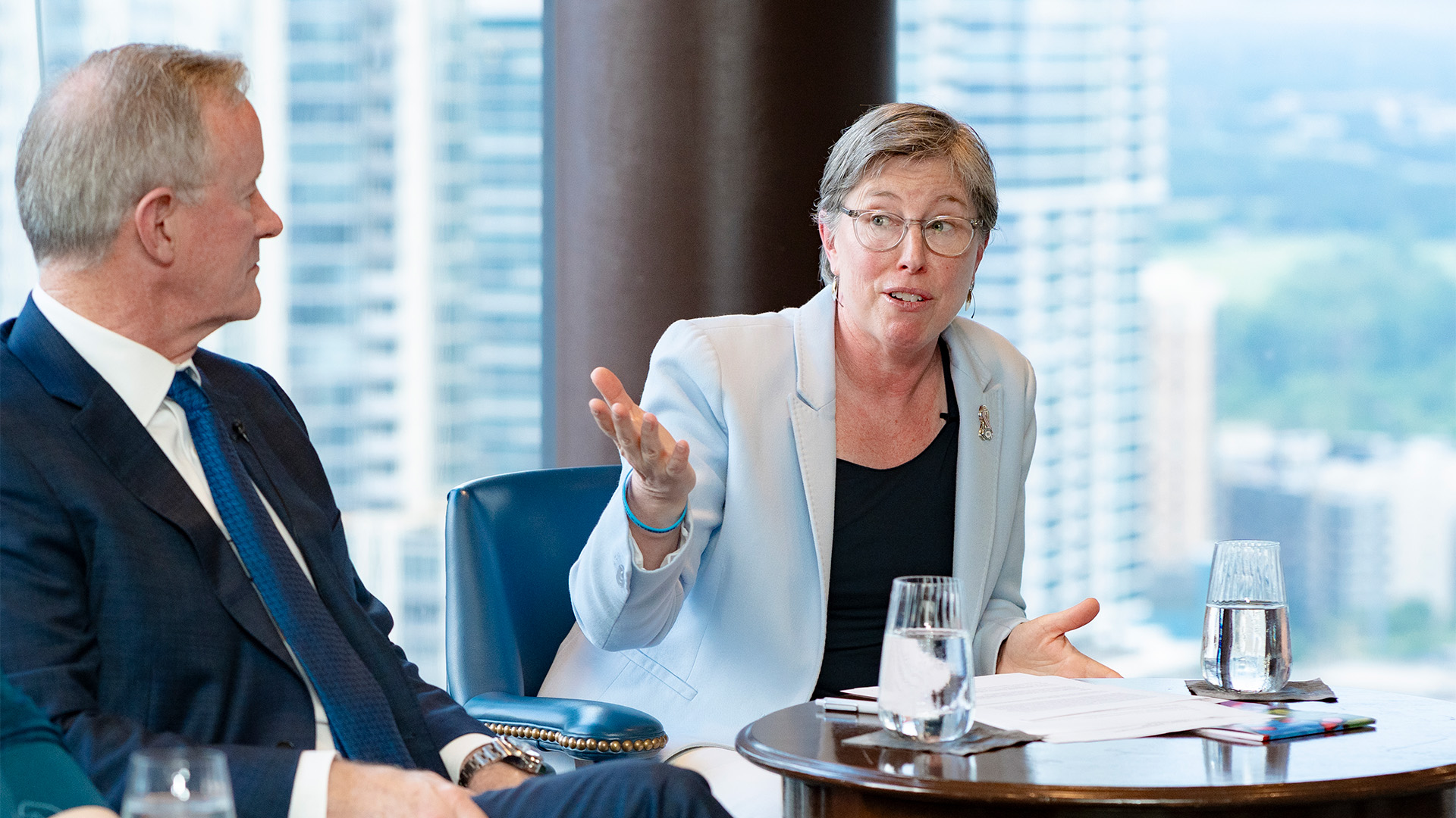 Two speakers, one woman and one man, seated in front of a view of the city out of the window behind them