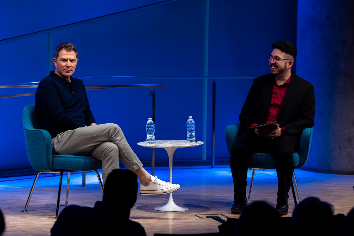 Bobby Flay and Alejandro Victoria sit on chairs on either side of a white round table with bottles of water on it