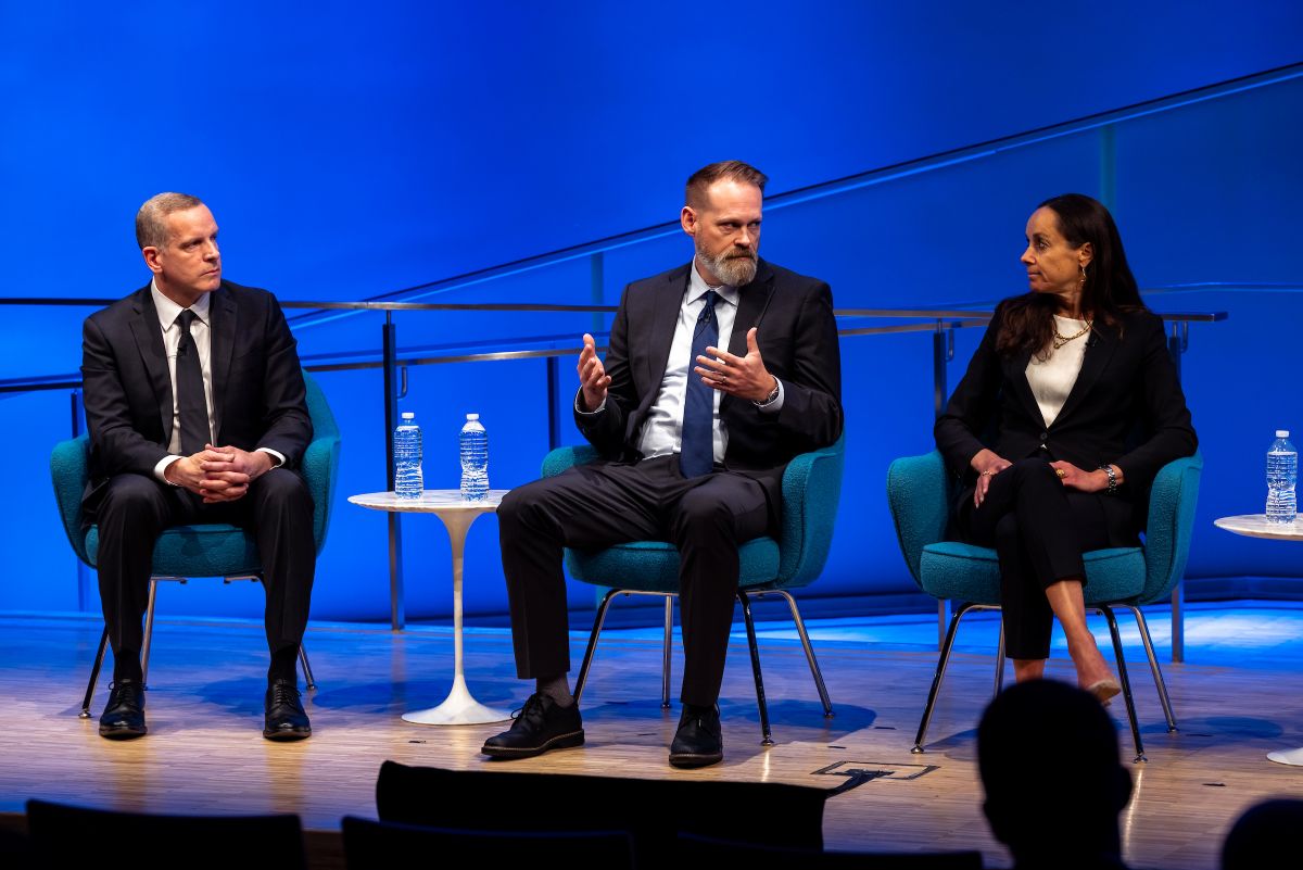 Three people talk on stage. A man in the middle gestures.
