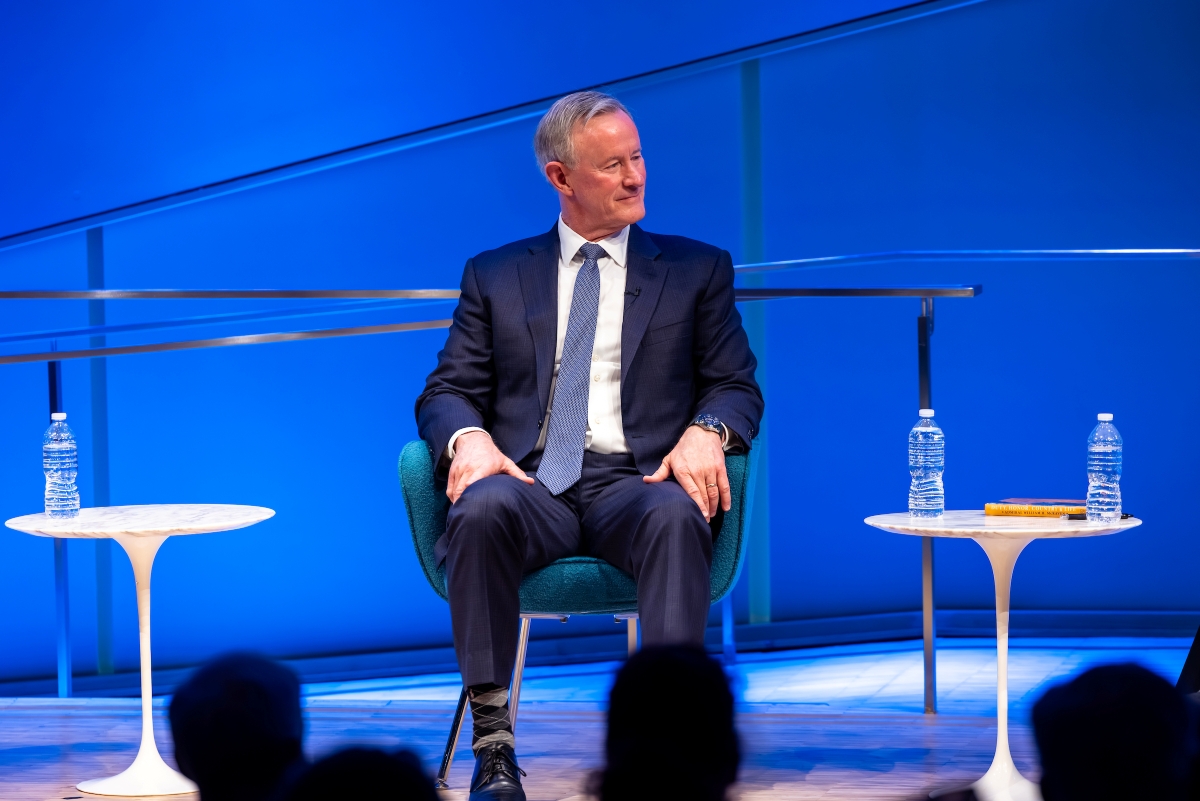 Admiral McRaven sits with his hands on his knees looking at others on the stage.