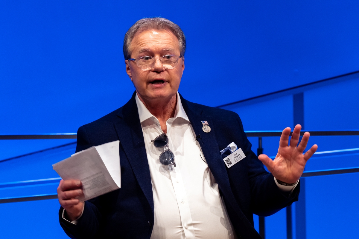 A man holds a folded piece of paper in his right hand and gestures with both hands, speaking.